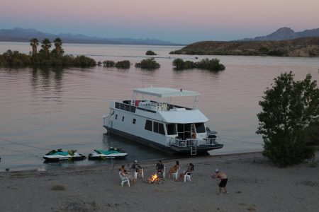 houseboat anchored at the beach shore with people sitting around a campfire on the beach | Cottonwood Cove Resort