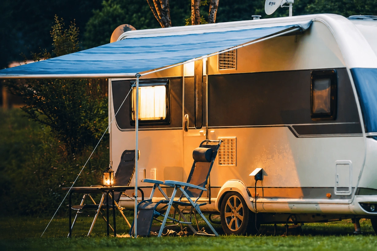 A white RV trailer parked at a campsite at dusk with its blue awning extended over two lawn chairs and a small table.